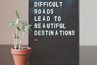 Difficult Roads Lead to Beautiful Destinations — a board bearing this inscription with a seedling in a pot next to it. Hope springs eternal.