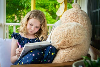 A young girl with a large teddy bear writing in a book