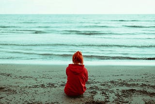 A person sits with their back to the camera in front of a tranquil sea. They have red hair in a ponytail and are wearing a red hooded sweatshirt.