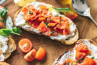 Two slice of bruschetta with cream cheese and tomato sit on a bread board next to a bottle of olive oil