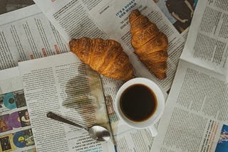 Cover image: Many newspapers with a 2 croissants, a spoon, a cup of coffee, and some coffee split over a newspaper.