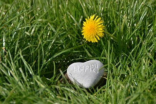 Photo of grass with a flowering dandelion, on the grass a heart-shaped rock that has the phrase “happy” engraved