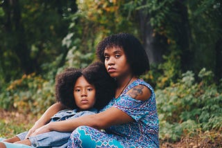 A Black mother and daughter are sitting on the ground in a forest. The daughter is leaning backwards on her mother. They both have serious looks on their faces.