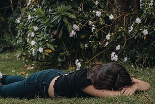 A woman wearing jeans and a black top lies face down in grass. A bush with flowers are behind her.