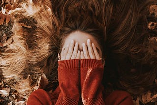 Girl on a floor of dry leaves with her hands over her face and wearing a red jumper