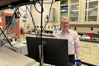 Brad Sutliff wears safety glasses as he stands in the lab in front of a computer monitor.