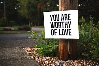 A big white sign that says, “You Are Worthy Of Love”, sits in front of a tree, behind which there is a green bush.