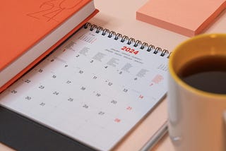 Paper calendar open to January 2024. It is on a desk with a pen, cup of coffee, and notepad