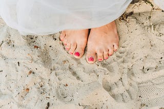 Pic of two feet with red-painted toenails partially covered by a white gown in the sand.