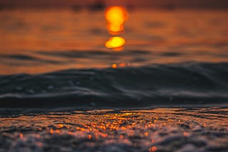 The image shows a vibrant sunset over the ocean. The sun, a glowing orange circle, is low on the horizon, casting a warm, golden reflection on the surface of the water. The waves are gently rippling, with the sunlight creating a shimmering path across them. The overall scene is serene and calming, with the soft, warm colors evoking a peaceful evening by the sea.