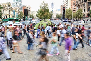 A large crowd of people crosses the road. Each of these people are a blurred rush as they busily cross the street.