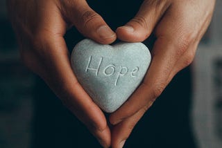 a gray stone heart with the word “hope” engraved held by two soft brown hands