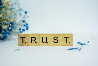 Scrabble game letter blocks spell “TRUST” on a white background with some small, blue and yellow flowers in one corner.