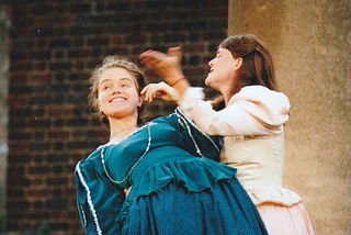 Rosalind and Celia in Shakespeare’s As You Like It. The actors are in Renaissance dresses. Rosalind lies playfully in Celia’s arms.