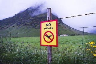 A barbed wire fence in front of a steep hill with some cloud cover at the top. The sign on the fence says “NO Drones.” There are some low buildings at the foot of the hill.