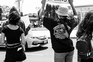 A black & white photograph of 4 people with backs turned, facing a street. One holds a sign that says “Remember their names.”