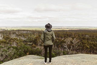 A woman wearing a hat and a green jacket is standing on mountain, looking at the sight below her.