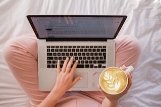 Woman sitting on her bed with her legs crossed drinking coffee and using her laptop.