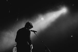 A man in dark clothes and a hoodie with his back to us in front of a microphone. It is dark, and smoky or cloudy, but two spotlights illuminate him,