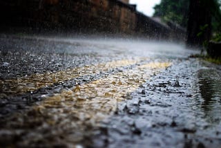 A picture of a road in the rain. The image is a close-up near the curb, over a double-yellow line. The rain is beating down heavily and the splashes are clearly visible. Blurred houses are in the background.