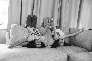 2 young girls lying upside-down on the back-support part of a sofa