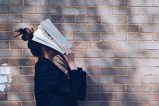 A young woman in front of a wall holds a text book over her face.