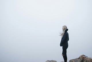 A woman standing on the edge of a rocky cliff, looking off into the foggy drop below