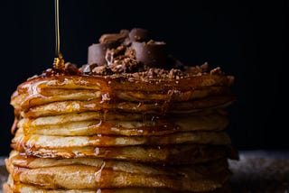 Pile of pancakes with syrup being poured, topped with chocolate pieces and shavings by Shay Wood