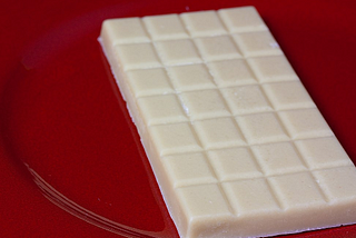 A photograph of a vegan white chocolate, scored in squares bar. It is placed on a red shiny circular surface. The background is blurred.