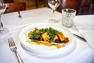 close-up on a fine dining table with white tablecloth and crystal wine and water glasses, and a white plate featuring a dish of greens and tofu with sauce, elegantly plated