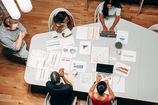 Cinco pessoas, em uma sala de reunião, ao redor de uma mesa, analisando informações em notebooks e vários papeis com gráficos e dados de pesquisas.