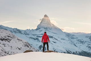 A man in a red coat on a snowy mountaintop looking at a sunrise.