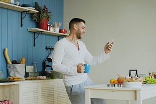 A smiling man in a kitchen looks at his smartphone while holding a blue mug.