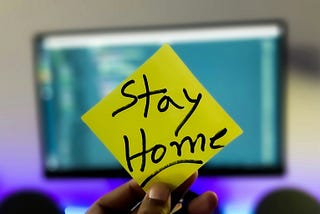 A hand holding a yellow sticker with the lettering: Stay Home.