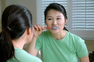 A light-skinned, middle-aged Asian woman brushes her teeth in a mirror. She has a full face of everyday makeup on and is wearing her straight black hair tied back into a ponytail with a few strands loose. She is wearing a mint green T-shirt. The woman appears to be in a 90s-style builder grade bathroom with taupe walls and white shutters visible behind her.