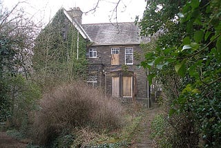 An abandoned, boarded up old house, surrounded by bushes and trees. Very spooky!