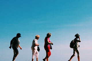 A group of elderly people walk ing. It seems like they are on the beach under a cloudless sky.