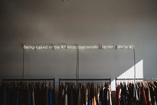 Neon lights above a clothing rack that reads: being naked is the #1 most sustainable option. we’re #2.