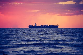 A cargo ship on the ocean, sat against a canvas sky of red and orange