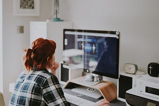 Caucasian, red-haired woman, with hair in ponytail pulled back, wearing blue and white plaid shirt facing her large computer monitor on raised platform, working at an engineering application, in what looks to be a home office.