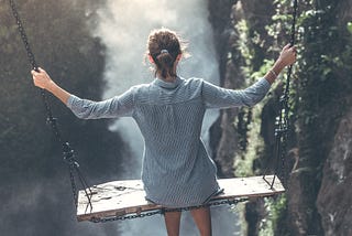 A picture of a lady on a swing swinging forward into a foggy narrow space between two steep cliffs. It signifies a jump into the unknown or murky waters but going anyway