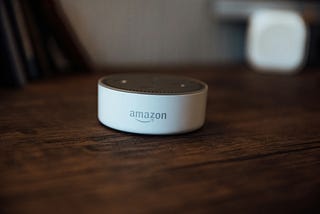 The image features an Amazon Echo Dot (3rd Generation) on a wooden surface. The device is compact and circular, primarily white with a gray top that includes speaker holes. In the background, there’s a slightly blurred white object, enhancing the focus on the Echo Dot.