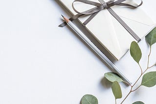envelope tied with a thin grey pink ribbon, silver pencil alongside, a sprig of greenery lying along the bottom, white background.