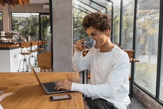 The photo of a short hair woman in a white long sleeve shirt drinking coffee while working on her laptop. ROMAN ODINTSOV/Pexels.