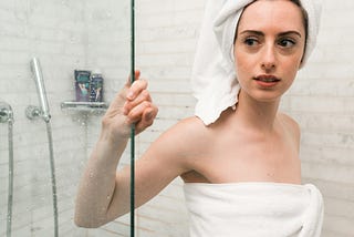 A white woman standing in a white-tiled shower. She has her hair wrapped in a white towel on top of her head, and her body is wrapped in another white towel. She is gripping the glass wall of the shower with her right hand.