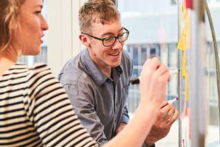 Two colleagues work together at a whiteboard, writing on several posted sticky notes.