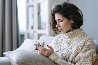 A depressed lady in a white knitted sweater is using a cell phone. Ivan Samkov/Pexels.