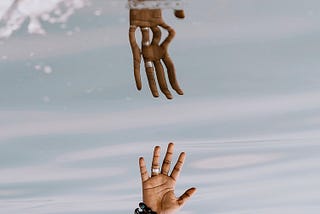 Two hands reach for each other. The one at the top is coming out of water to grasp the hand coming from the below.