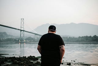 Man standing at a river’s edge pondering, bridge and mountains in the distance