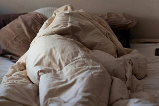 Someone wrapped up in tan sheets. They appear to be so cozy that they've even covered up their heads. Its summer, there is sunlight coming to the window shining onto the blankets.